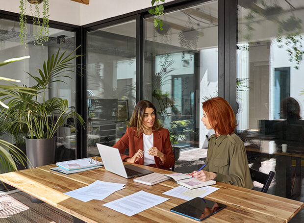 Two busy business women of young and middle age talking in creative green office sitting at desk. Professional ladies employee and manager having conversation using laptop at work. Candid photo.